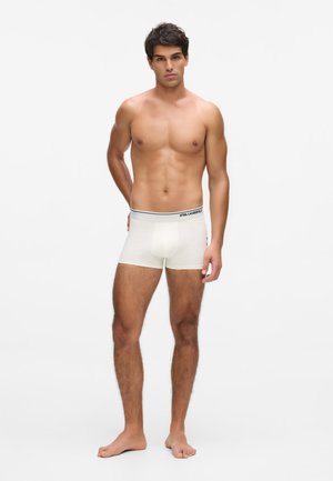 Young man standing barefoot, shirtless, wearing white boxer briefs with "Karl Lagerfeld" logo on waistband against a plain white background.