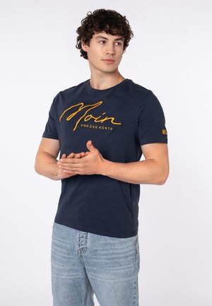 Junger Mann mit lockigem Haar, der ein dunkelblaues T-Shirt mit orangefarbenem Schriftzug trägt, steht mit verschränkten Händen und schaut zur Seite.