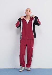 Bordeaux tracksuit med sorte og hvide accenter, zip-up jakke og bukser med elastisk talje. Sølvfarvede træningssko fuldender outfittet.