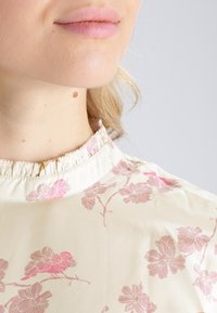 Ljus krämfärgad blus med rosa blommönster, rynkad krage och slät textur. Närbild av halsområdet och blommönstret.