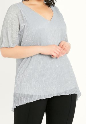 Woman wearing a light gray pleated short-sleeve top with a V-neck, paired with black pants, hands clasped in front.