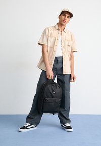 Camisa bege de mangas curtas com botões, top branco, calças cinzas de perna larga, mochila preta e ténis preto e branco. Traços de design simples.