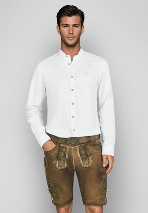 Mann trägt traditionelle braune Lederhose mit Stickereien und ein weißes Hemd mit Knöpfen, steht vor einem schlichten grauen Hintergrund.