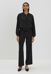 Schwarze, kurze Jacke mit Stehkragen und elastischem Bund, kombiniert mit schwarzen, hoch geschnittenen, weiten Hosen, beide aus glattem Material.