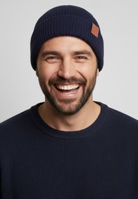 Navy ribbed beanie with a textured finish and a leather label. Worn by a man in a matching navy textured sweater, smiling.