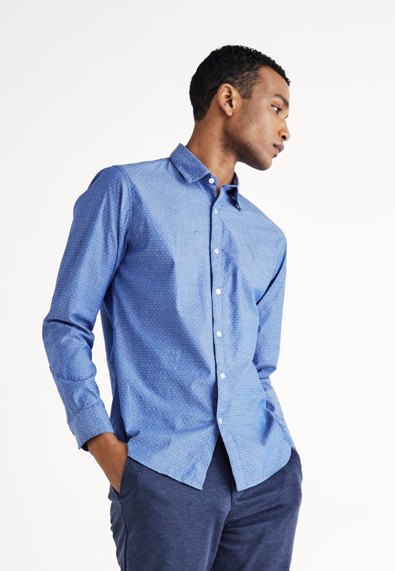Camisa de manga larga azul claro con un patrón sutil, cuello abotonado y botones en la parte frontal. La tela parece suave y ligeramente texturizada.
