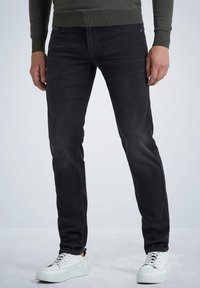Zwarte denim spijkerbroek, slim fit, met vijf zakken en een verweerde afwerking. Gecombineerd met witte sneakers met textuur en een donkergroene trui.