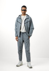 Pelēks sporta jaka ar melniem akcentiem un rāvējslēdzēja kabatām, kombinēta ar atbilstošām pelēkām biksēm. Balti sporta apavi noslēdz izskatu.