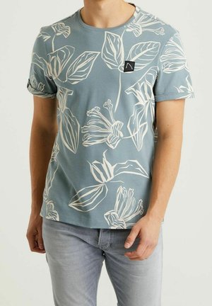 Homme portant un t-shirt bleu à manches courtes avec de grands imprimés floraux blancs et un jean gris clair, debout avec une main près de la poche.