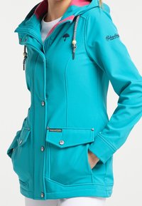 Kvinna i en turkos hoodie-jacka med snören, framfickor, tryckknappar och en liten paraplylogotyp på bröstet.