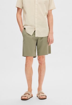 Pantalones cortos verde oliva con bolsillos laterales, combinados con una camisa de manga corta beige claro y sandalias beige abiertas con tiras.