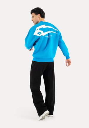 Mann trägt blauen Pullover mit weißem Motiv auf dem Rücken, schwarze Hose und weiße Sneakers, steht mit dem Rücken zur Kamera und schaut nach links.