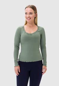 Maglietta a maniche lunghe di colore verde chiaro con una texture a coste, bordo in pizzo lungo il colletto e bordi ondulati; abbinata a leggings scuri.