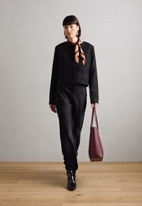 Zwarte cropped jack met twee voorzakken, gecombineerd met een zwarte jurk, glanzende zwarte laarzen en een bordeauxrode tote bag. Satijnen sjaal om de nek.
