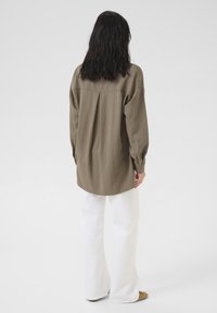 Olive green oversized shirt with long sleeves, a back yoke seam, and button cuffs, paired with white wide-leg pants and beige footwear.