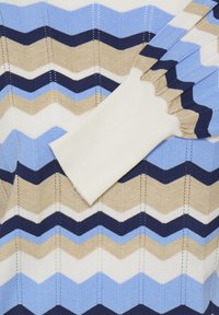 Pull en tricot présentant un motif en zigzag bleu clair, bleu foncé, beige et blanc avec des poignets côtelés blancs. Tissu texturé avec des rayures verticales.