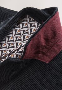 Nahaufnahme eines schwarzen Cordkragens mit gemustertem Innenfutter in Weiß, Schwarz und Orange und einem bordeauxroten Filz-Unterkragen.