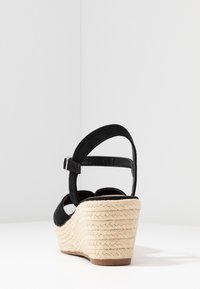 Sandale compensée en suède noir avec une semelle en jute tissée, bride de cheville ajustable et design à bout arrondi. La texture est lisse sur la tige.