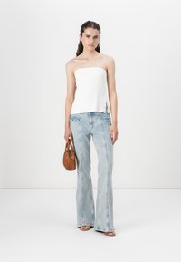 Hvit stroppeløs topp, lyseblå utsvingte jeans med sidelommer, og en brun håndveske. Modellen har på seg lyse sandaler og står mot en hvit bakgrunn.