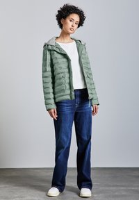 Ljusgrön pufferjacka med grå huva, dragkedjefickor och en slät yta, tillsammans med mörkblå jeans och vita sneakers.