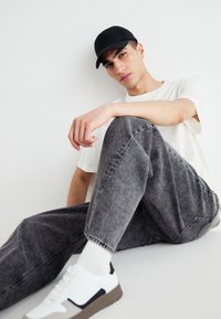 Black cap, white t-shirt, and gray denim jeans. Casual white sneakers with gray and navy accents. Subject sitting against a white background.