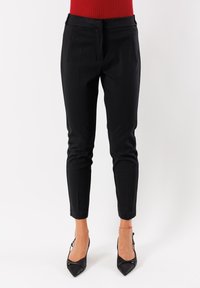 Donna che indossa pantaloni neri slim alla caviglia e tacchi a punta neri, in piedi contro uno sfondo bianco.