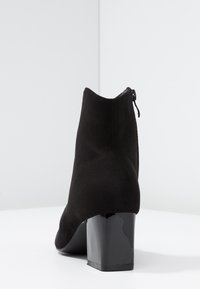 Bota de tobillo de ante negro con una textura suave, punta redondeada y un tacón grueso y brillante. El detalle del zipper en la parte trasera mejora la funcionalidad.