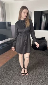 Rochie neagră cu nasturi, mâneci lungi, talie strâmtă, fustă evazată. Asortată cu sandale negre cu bretele. Podea din lemn deschis la culoare, mobilier închis la culoare în fundal.