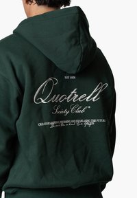 Dunkelgrüner Hoodie mit Kapuze und Bindebändern. Auf der Rückseite befindet sich großer silberner Text: "Quotrell Society Club" und ein zusätzlicher Slogan darunter.