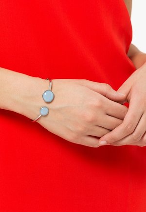 Mains jointes au-dessus d'un poignet portant un haut rouge et un bracelet ouvert en argent avec deux pierres rondes bleu pâle.