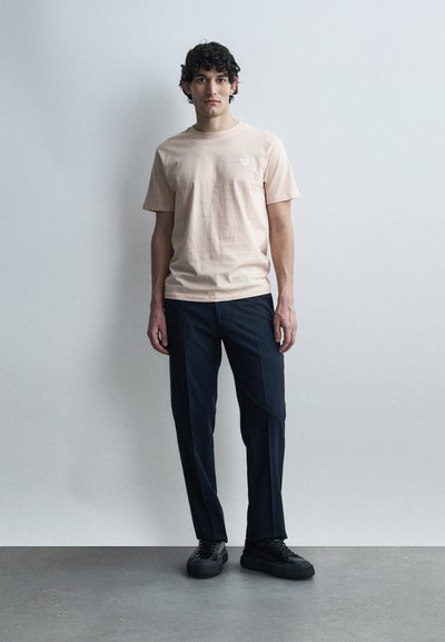 Άνδρας με σγουρά μαλλιά στέκεται μπροστά σε ανοιχτό γκρι τοίχο φορώντας ανοιχτό μπεζ κοντομάνικο πουκάμισο, σκούρο μπλε παντελόνι και μαύρα αθλητικά παπούτσια.