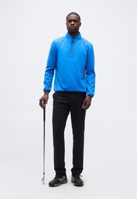 Άνδρας στέκεται κρατώντας μπαστούνι γκολφ, φορώντας φωτεινό μπλε πουλόβερ με φερμουάρ και μαύρο παντελόνι, στο φόντο απλού λευκού.