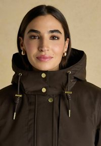 Hnedá kapucňa s kovovým zlatým príslušenstvom, patentnými gombíkmi a šnúrkami. Tkanina je hladká s lesklým povrchom a má vysoký golier.