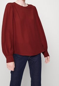 Burgundy pitkähihaiset paita, jossa on pyöreä kaula-aukko, ryGatheroudu sitaatankain reunoissa ja istuvat hihansuut, yhdistettyinä tummiin farkkuihin. Pehmeä, sileä kangas.