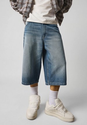 Persoon die lichtblauwe, wijde denim shorts draagt, witte sokken, witte sneakers, een beige T-shirt en een bruine geruite overshirt tegen een effen achtergrond.