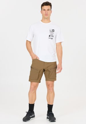 Mann trägt weißes T-Shirt mit Grafik, braune Cargo-Shorts mit Reißverschlusstaschen, schwarze Socken und schwarze Sportschuhe, steht mit einer Hand in der Tasche.