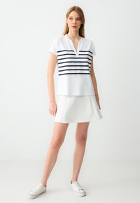 Weißes Polohemd mit navyblauen horizontalen Streifen, kurzen Ärmeln und V-Ausschnitt. Kombiniert mit einem weißen Rock und weißen Turnschuhen vor einem schlichten Hintergrund.