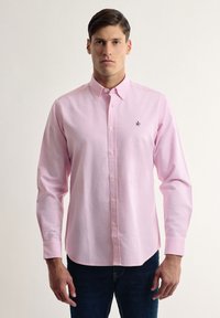 Camisa de botones rosa claro hecha de algod�ón, con mangas largas, cuello y un pequeño logo oscuro en el lado izquierdo del pecho. Llevada con jeans oscuros.