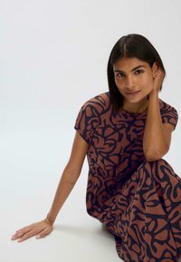 Ruskea mekko mustilla abstrakteilla kuvioilla, lyhyt hihaisena ja virtaavana siluetilla. Sileä kangas, istuma-asennossa, käsi lepotilassa jalalla.