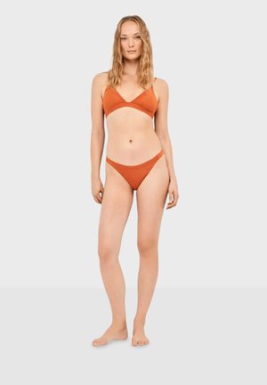 Žena stoji bosonoga noseći teksturirani bikini boje izgorenog narančastog, s trokutastim gornjim dijelom i niskim donjim dijelom, na jednobojnoj svijetloj pozadini.