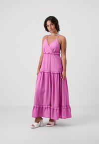 Rochie maxi de culoare roz aprins, cu un design înfășurat, accente volănașe la talie și la margine, realizată dintr-un material ușor. Asortată cu sandale cu toc albe.