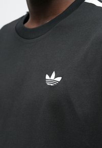 Μαύρο βαμβακερό T-shirt με ριμπ λαιμόκοψη, λευκό κεντημένο λογότυπο στο στήθος και λευκές ρίγες στους ώμους.