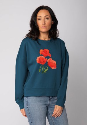 Femme aux cheveux bruns portant un sweatshirt teal avec trois fleurs de pavot rouges et un jean bleu devant un fond clair uni.