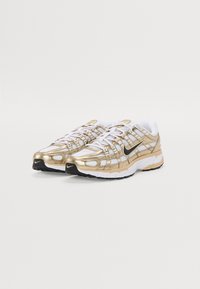 Zlatne i bijele atletske tenisice s mrežastim gornjištem, metalnim detaljima, uzorkom u obliku mreže i crnim potplatom. Nike logo na strani.