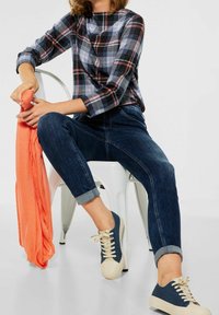 Nainen istuu valkoisella metallituolilla, pukeutuneena sinivihreään ruutupaitaan, käännettyihin tummansinisiin farkkuihin ja laivastonsinisiin lenkkareihin, pitäen käsissään korallinväristä huivia.