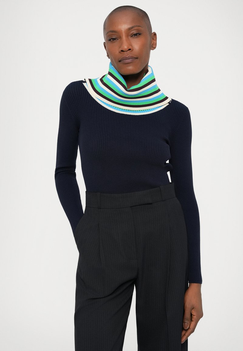 Navy ribbed longsleeve top met een meerkleurige gestreepte hoge kraag; gecombineerd met zwarte pinstripe broek.