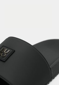 Sandale negre cu talpă plată, având un baretă lată și netedă, prezentând un suport pentru picior texturat și un mic patch cu logo embosat.