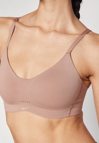Lys rosa sports-bh med justerbare stropper, glatt tekstur, subtilt Nike-logo, og perforerte detaljer på midten foran.