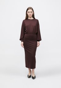 Bordowa sukienka o luźnym kroju, z długimi rękawami i marszczonym paskiem w talii, zestawiona z czarnymi butami z szpiczastym noskiem. Gładka faktura, długość do kostek.