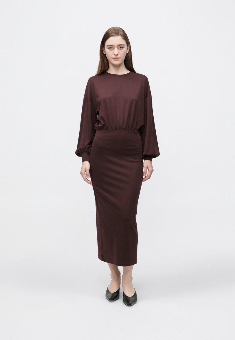 Bordowa sukienka o luźnym kroju, z długimi rękawami i marszczonym paskiem w talii, zestawiona z czarnymi butami z szpiczastym noskiem. Gładka faktura, długość do kostek.